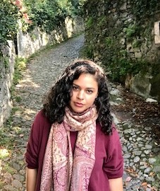 A brunette woman with curly hair, a burgundy scarf, and a cobblestoned path with moss in the background.