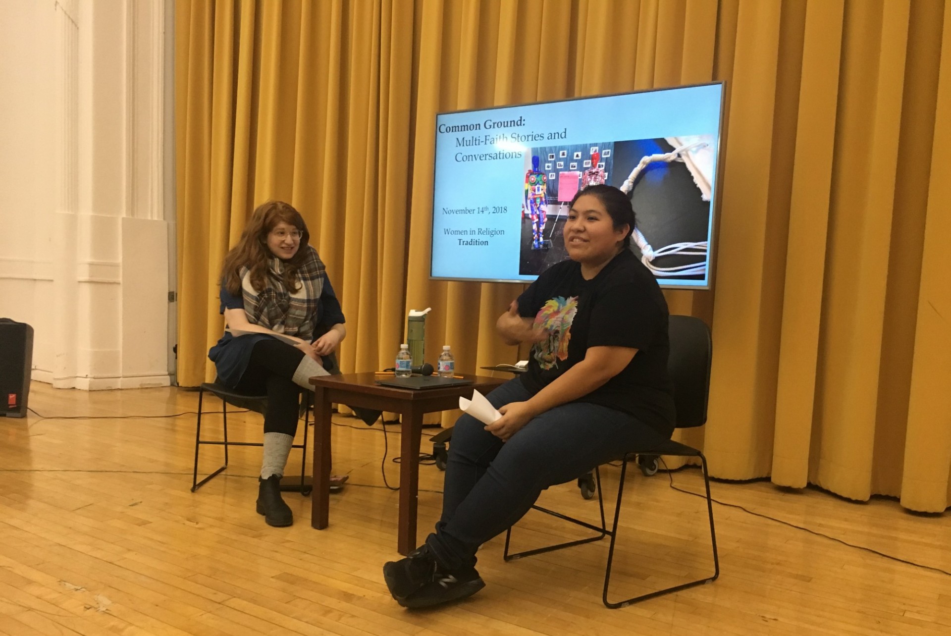 Jessica Tezen and Atara Cohen speaking at a Common Ground Event in the Earl Hall Auditorium in front of the mustard curtain.