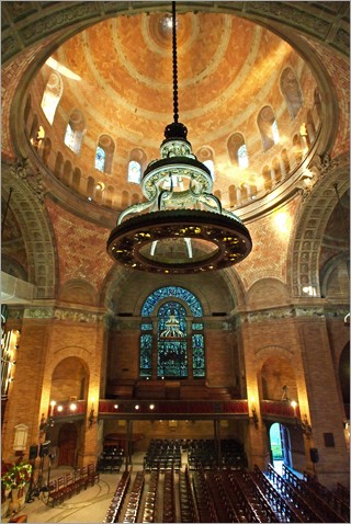 South facing view of the interior of the chapel that includes sun streaming in through the dome windows, the chandelier lit, seating, and the south transept. 