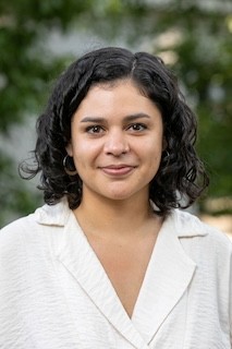 Brown-skinned woman with dark brown ringlets and small gold hoop earrings. Cream-colored top and blurred green trees in the background.
